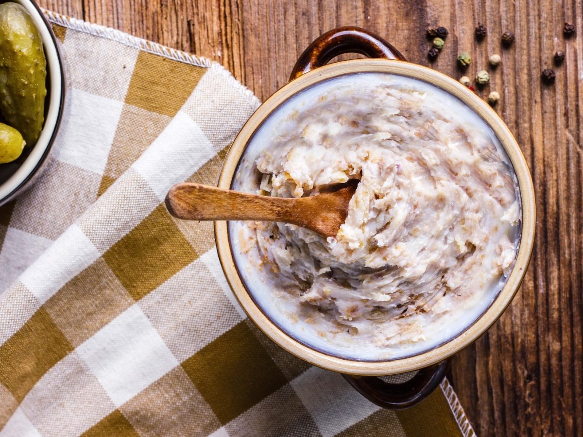 Veganes Zwiebelschmalz in einem Glas mit einem Löffel. Links davon ein Geschirrtuch und ein Glas mit sauren Gurken. Der Hintergrund ist hölzern.