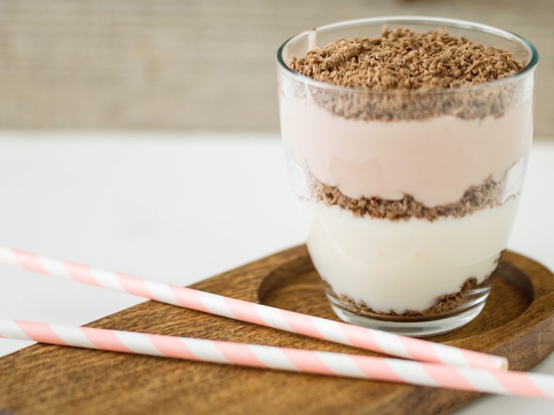 Ein Glas mit dem Dessert und zwei rosa-weißen-Strohhalmen auf einem braunen Untersetzer und hellem Untergrund.