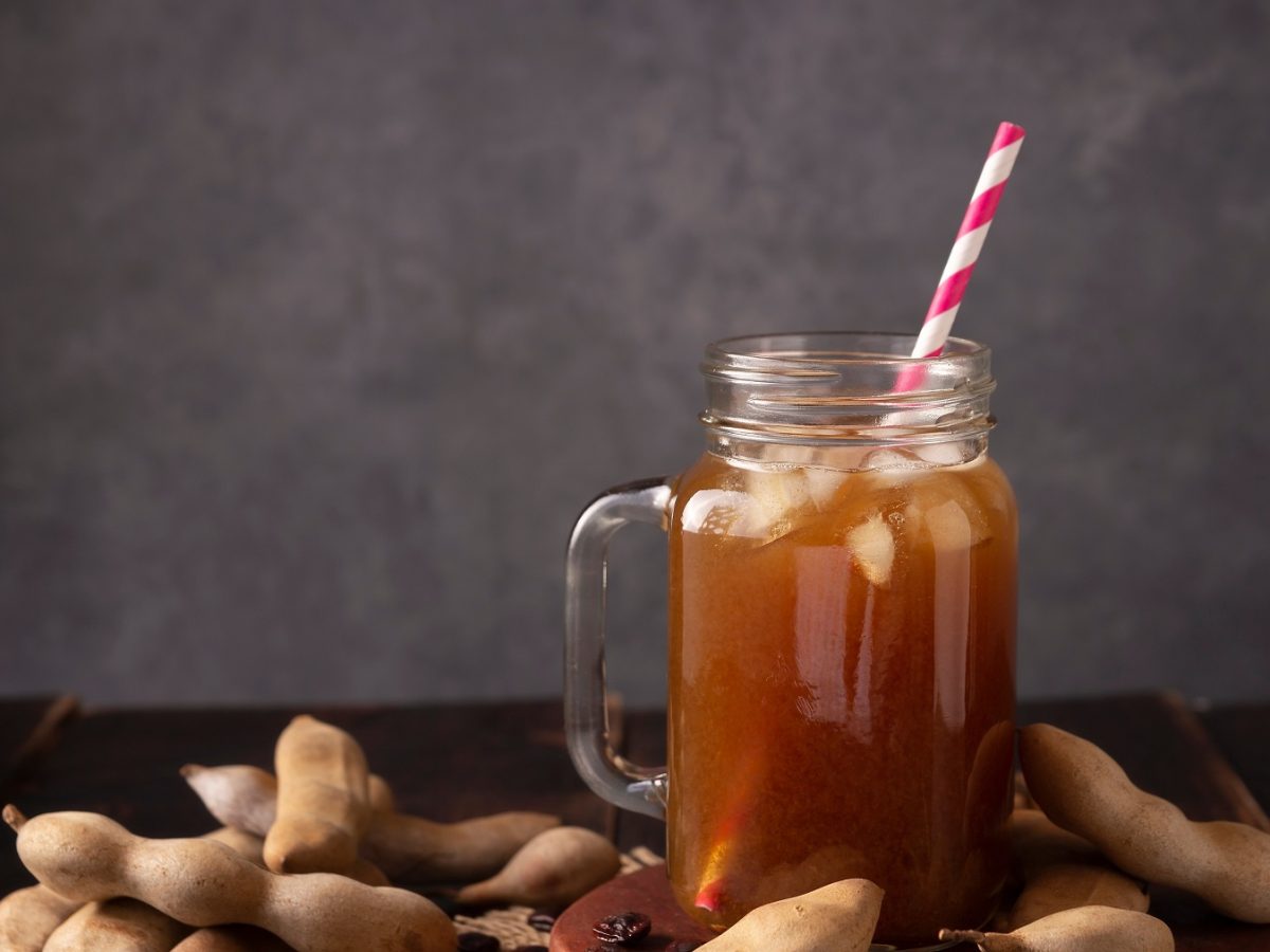 Agua de tamarindo in einem Glas mit henkel und einem gestreiften Strohhalm vor dunklem Hintergrund, daneben Tamrindenschoten.