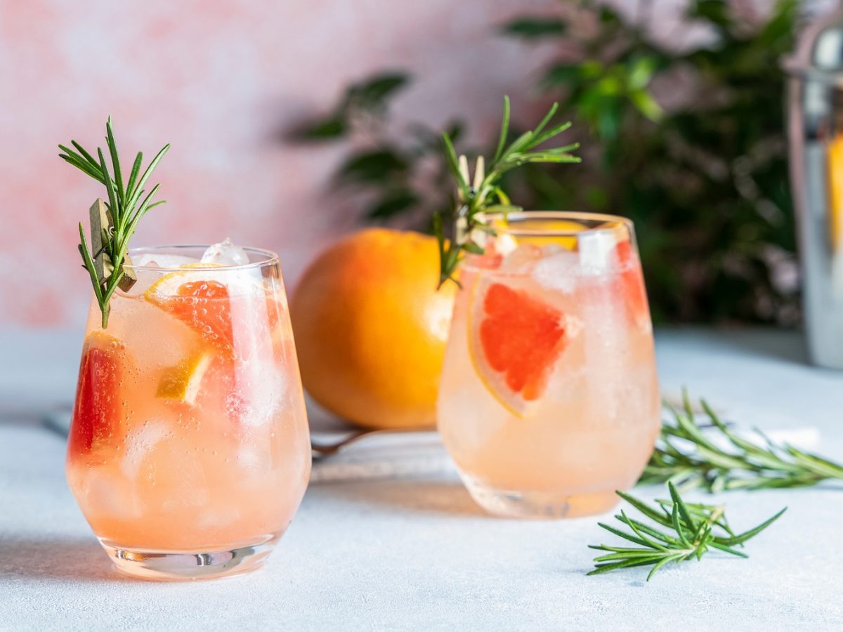 Zwei Gläser mit dem Amalfi Spritz auf einem hellen Tisch mit rötlichem Hintergrund. Alles von der Seite fotografiert und im Hintergrund noch Bar-Equipment und Pflanzen erkennbar.