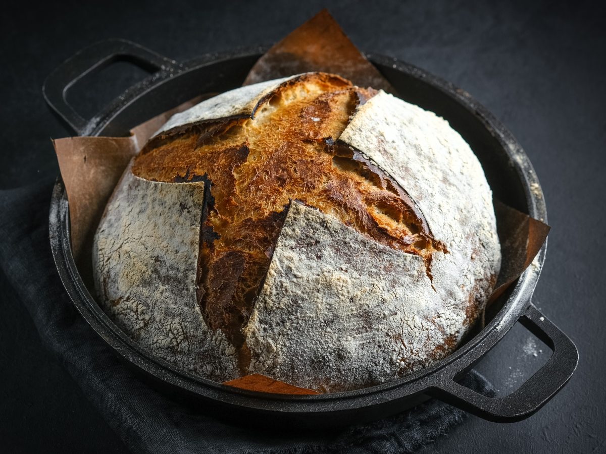 Ein gusseiserner Topf mit dem Brot auf einem schwarzen Tisch mit grauem Tuch. Alles von schräg oben fotografiert.