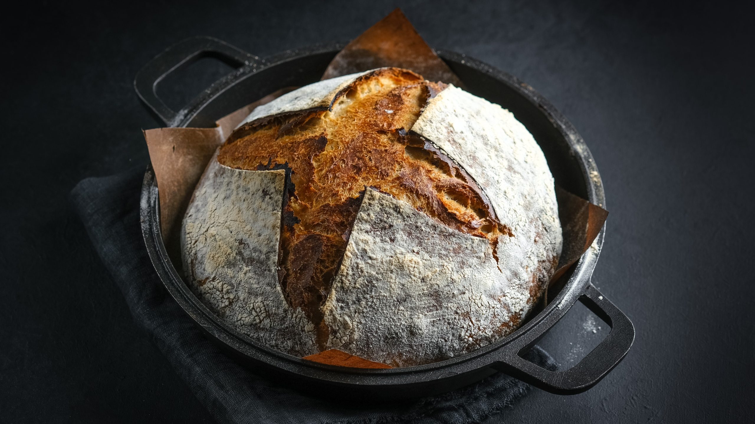 Ein gusseiserner Topf mit dem Brot auf einem schwarzen Tisch mit grauem Tuch. Alles von schräg oben fotografiert.
