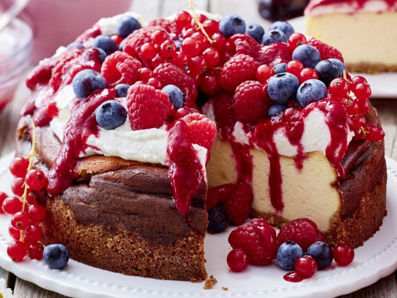 Beeren-Käsekuchen auf einem weißen Teller auf hölzernem Hintergrund. Hinten auf dem Bild ein Stück Beeren-Käsekuchen.