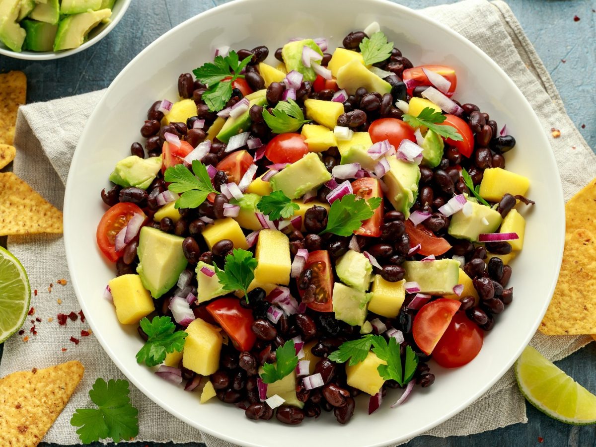 Bohnensalat mit Avocado, Mango, Tomaten, roten Zwiebeln und Mango in einer weißen Schüssel. Daneben Tortillachips und Limette.