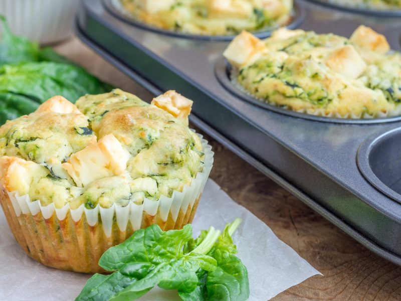 Mehrere Brokkoli-Feta-Muffins auf einem Holztisch und einige noch in der Backform dahinter. Alles von der Seite fotografiert, mit frischem Spinat.