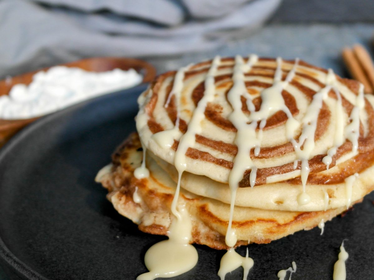 Mehrere Pfannkuchen mit Zimt-Twist auf einem dunklen Teller und grauem Tisch mit Zimt und Mehl im Hintergrund.