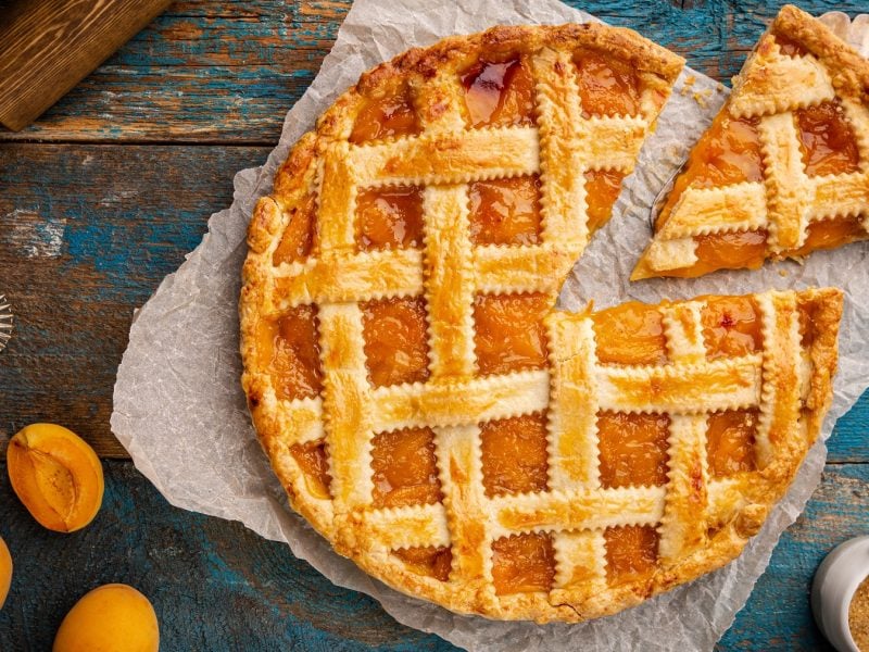 Goldbraune Crostata all'albicocca auf Backpapier mit einem Stück im Vordergrund, umgeben von frischen Aprikosen und Backutensilien.