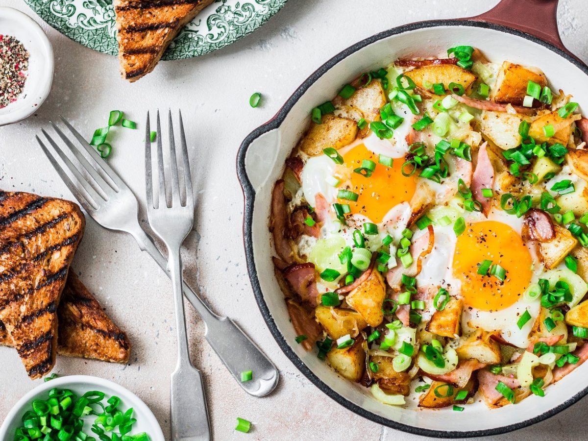 Draufsicht: Eine helle Pfanne mit einem deftigen Bauernfrühstück steht auf einem hellen Untergrund aus Holz. Drum herum liegen frische Toasts, Besteck sowie frischer Schnittlauch.