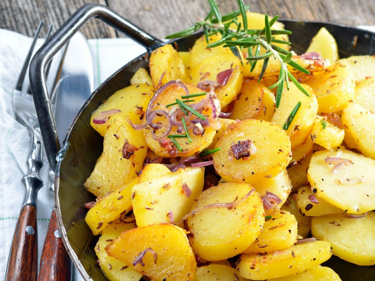 Draufsicht, schräg: Eine Pfanne mit Bratkartoffeln aus gekochten Kartoffeln. Die steht auf einem Brettchen, dass auf einem Tuch und das auf einem Holzboden.