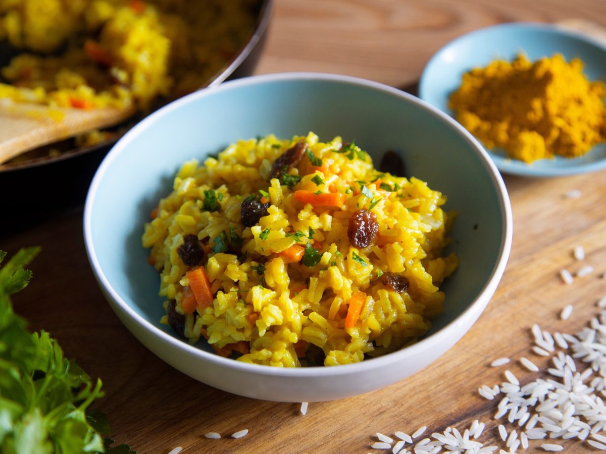 Draufsicht, schräg: eine hellblaue Schale Curry-Reis-Salat mit Rosinen auf einem Holzbrettchen. Daneben steht eine kleine hellblaue Schale mit Currypulver.