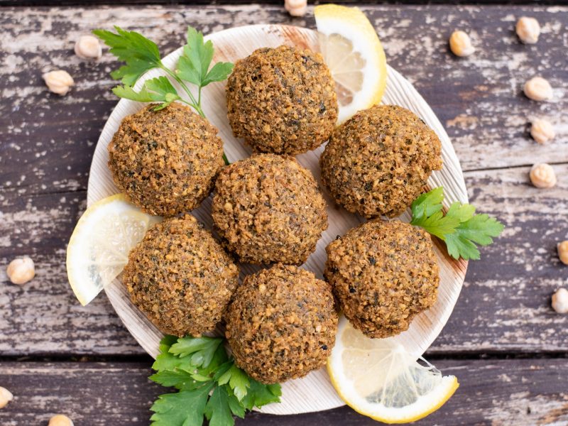 Draufsicht: Ein Teller mit Falafel aus dem Backofen steht auf einem rustikalen Holzboden. Drum herum liegen Kichererbsen.