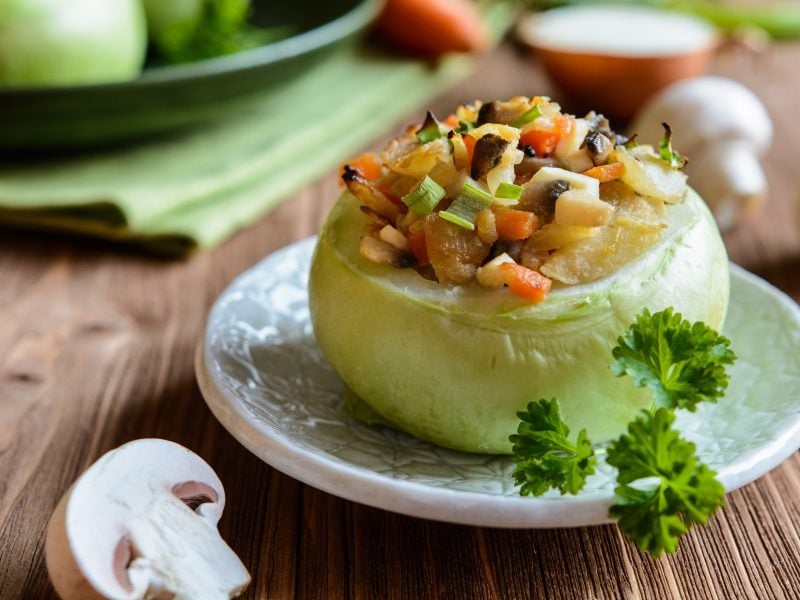 Draufsicht, schräg: eine gefüllte Kohlrabi mit Champignons steht auf einem Untergrund aus Holz. Drum herum liegen frische Pilze und Gemüse.