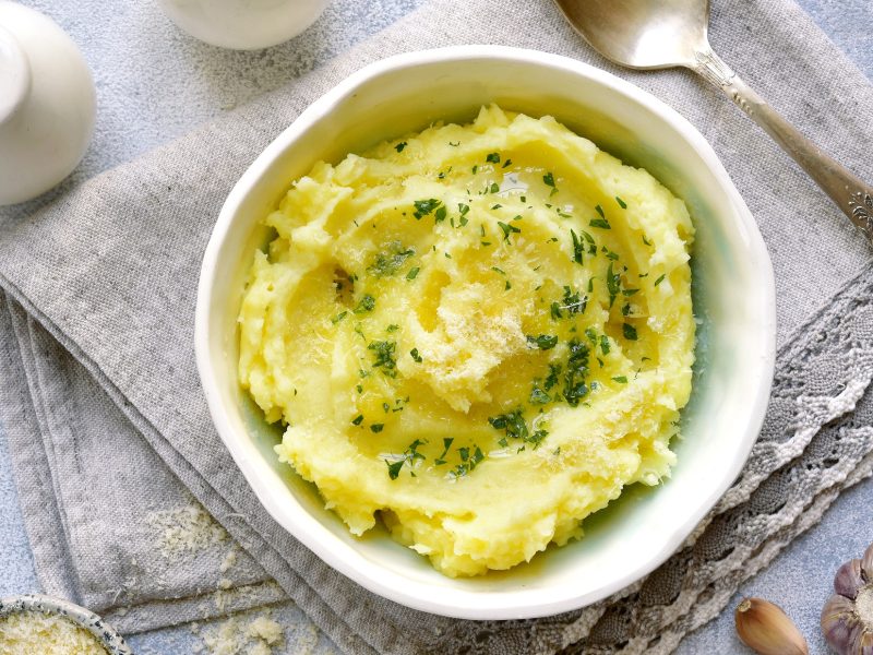 Draufsicht: eine weiße Schüssel Kartoffel-Thymian-Püree mit Käse auf einem hellblauen Untergrund aus Stoff. Daneben liegen ein silberner Löffel, ein Salzstreuer, ein Schälchen mit geriebenen Käse und ein Lorbeerblatt,