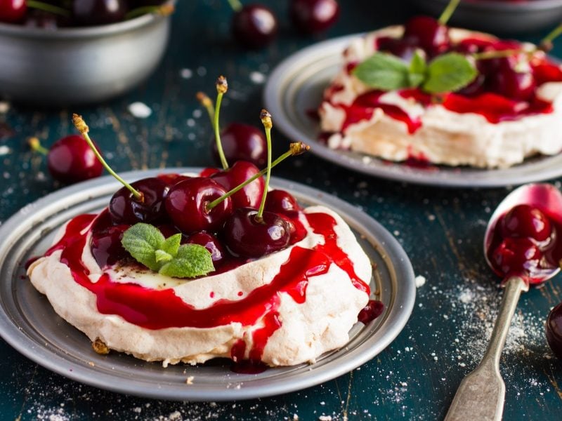 Draufsicht, schräg: Zwei Teller aus Metall mit je einer Kirsch-Pavlova. Drum herum liegen frische Kirschen und Besteck.