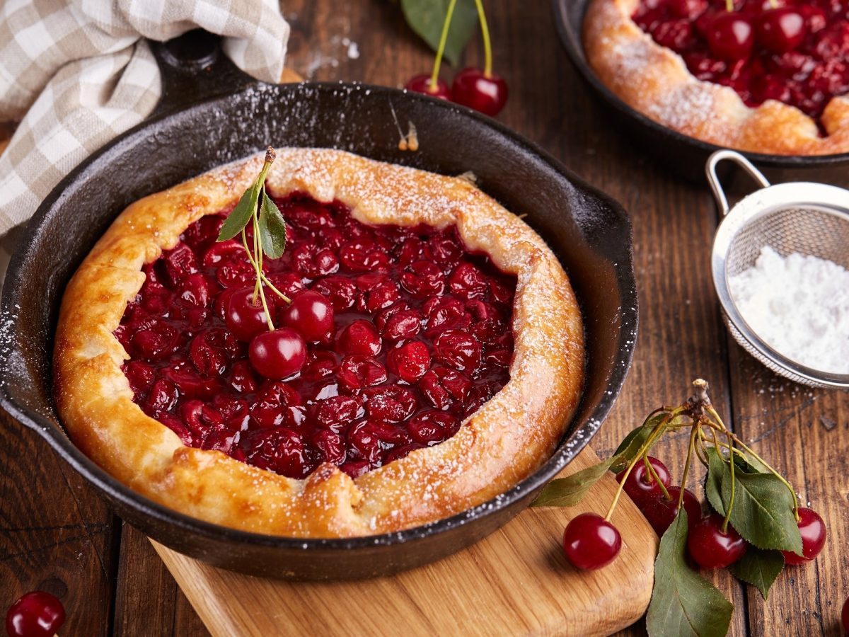 Draufsicht: Zwei Kirschen-Galette in einer ofenfesten. dunklen Form liegen auf einemr rustikalen Holzoberfläche. Drum herum sind lose Kirschen und ein weißes Tuch.