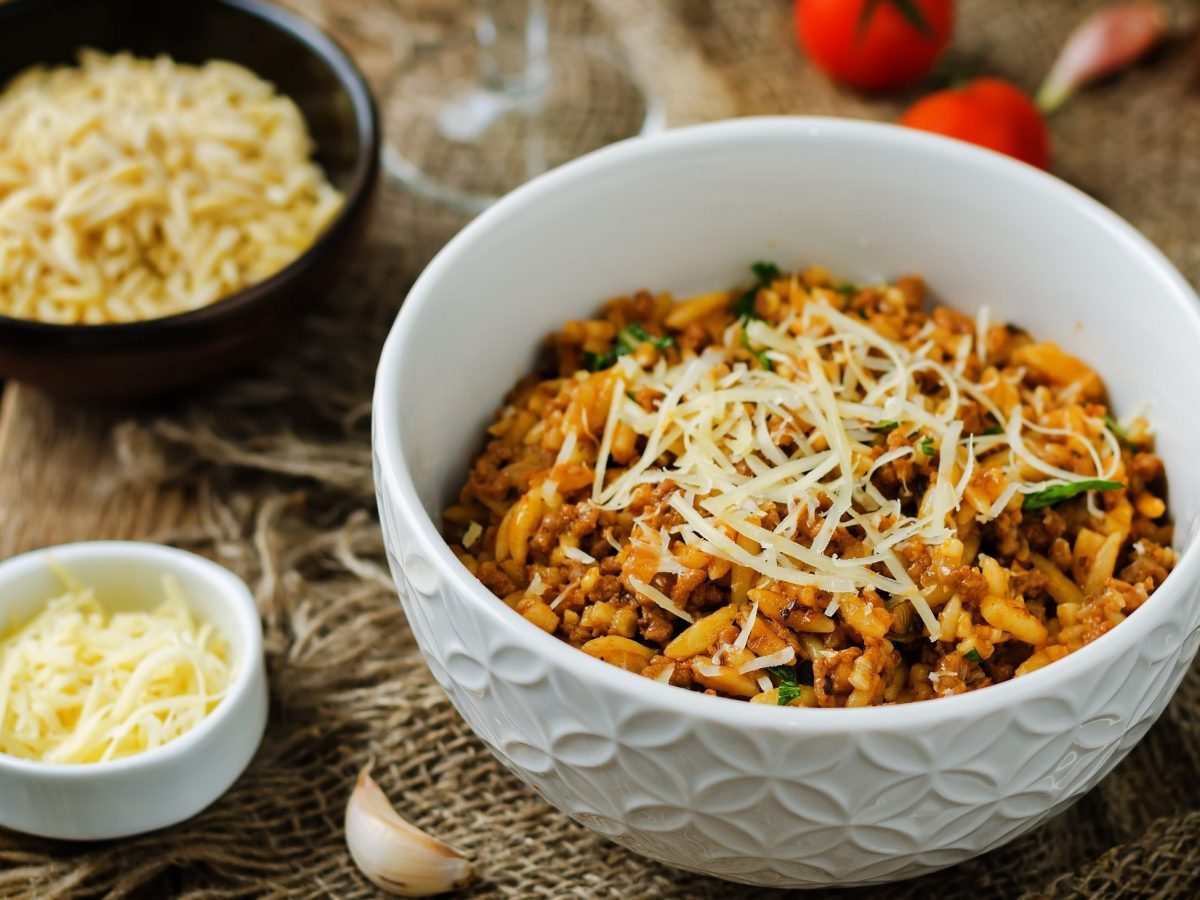 Draufsicht, schräg: Auf einem Untergrund aus Holz stehen drei Schüsseln. Eine große mit Kritharaki-Salat mit Hackfleisch. Und zwei kleine mit geriebenen Käse. Drum herum liegen einige frische Tomaten.