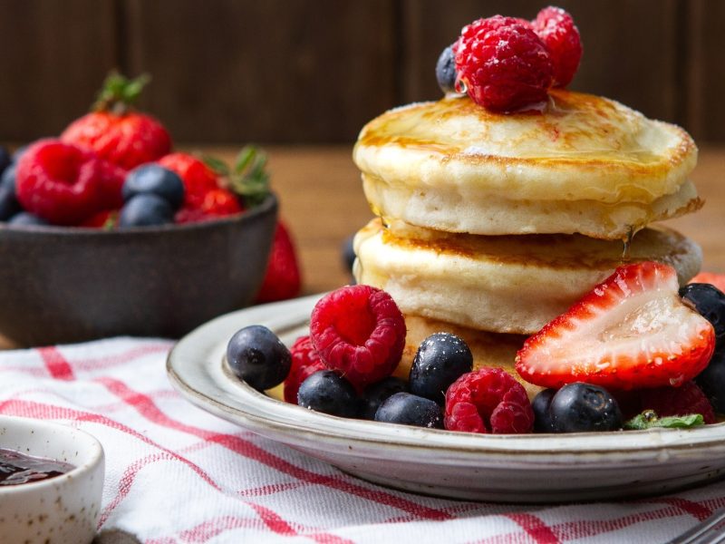 Frontal: Drei luffy Pancakes liegen gestapelt auf einem weißen Teller. Drum herum und auf den Pfannkuchen liegen frische Beeren.