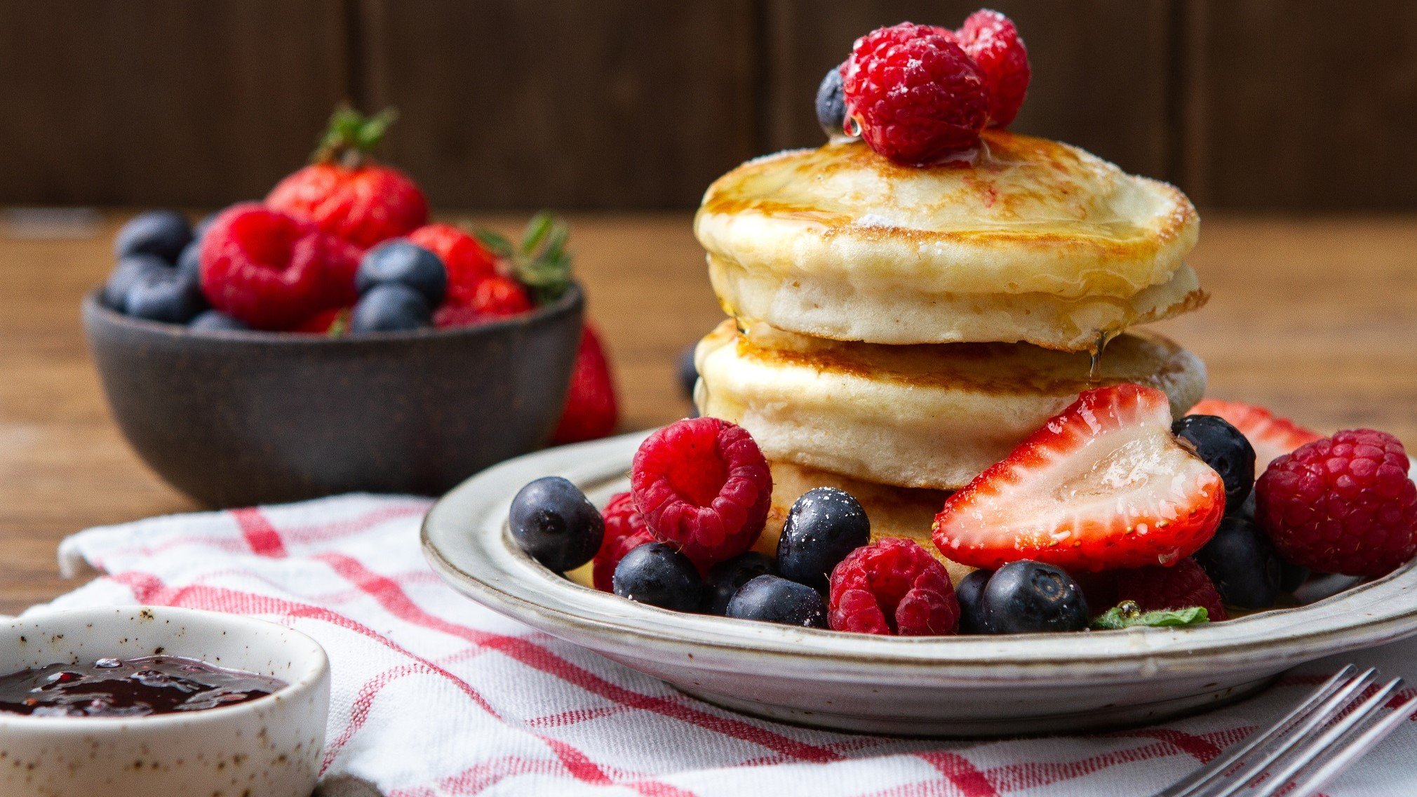 Frontal: Drei luffy Pancakes liegen gestapelt auf einem weißen Teller. Drum herum und auf den Pfannkuchen liegen frische Beeren.