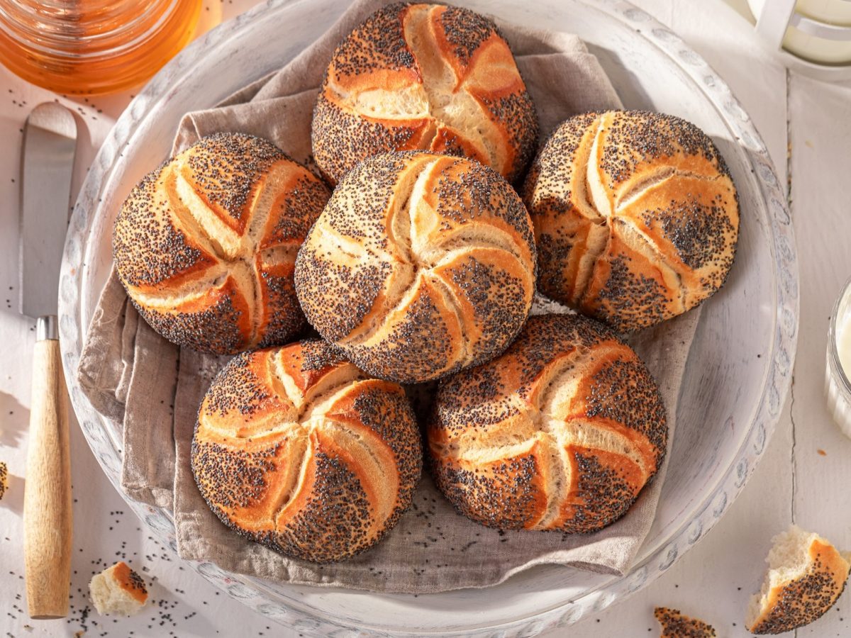 Draufsicht: Ein Frühstückstisch mit selbst gebackenen Mohnbrötchen aus Hefeteig. Drum herum sind ein weißes Schälchen mit Butter und und ein Glas Honig.