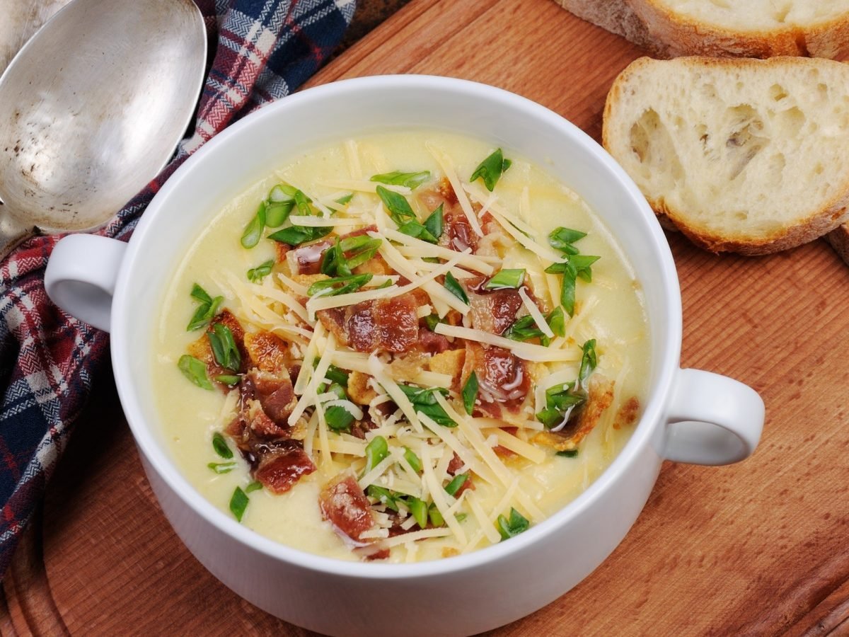 Draufsicht: Eine Pommersche Kartoffelsuppe in einer Suppentasse auf einem Untergrund aus Holz. Daneben liegen Weißbrotscheiben und zwei silberne Löffel.