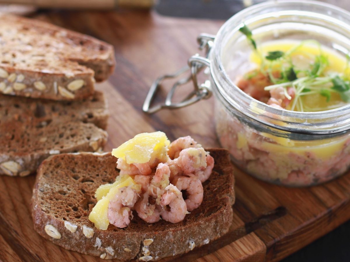 Ein Weckglas mit Potted Schrimps steht auf einem Holzbrettchen. Drum herum liegen mehrere Brotscheiben.