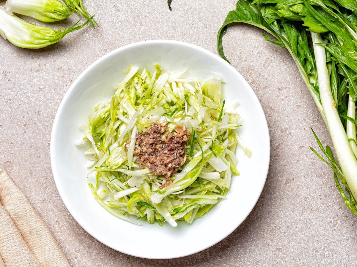 Draufsicht: Ein Teller Puntarelle-Salat mit Sardellen auf hellem Untergrund. Drum herum liegen Puntarelle-Stängel.