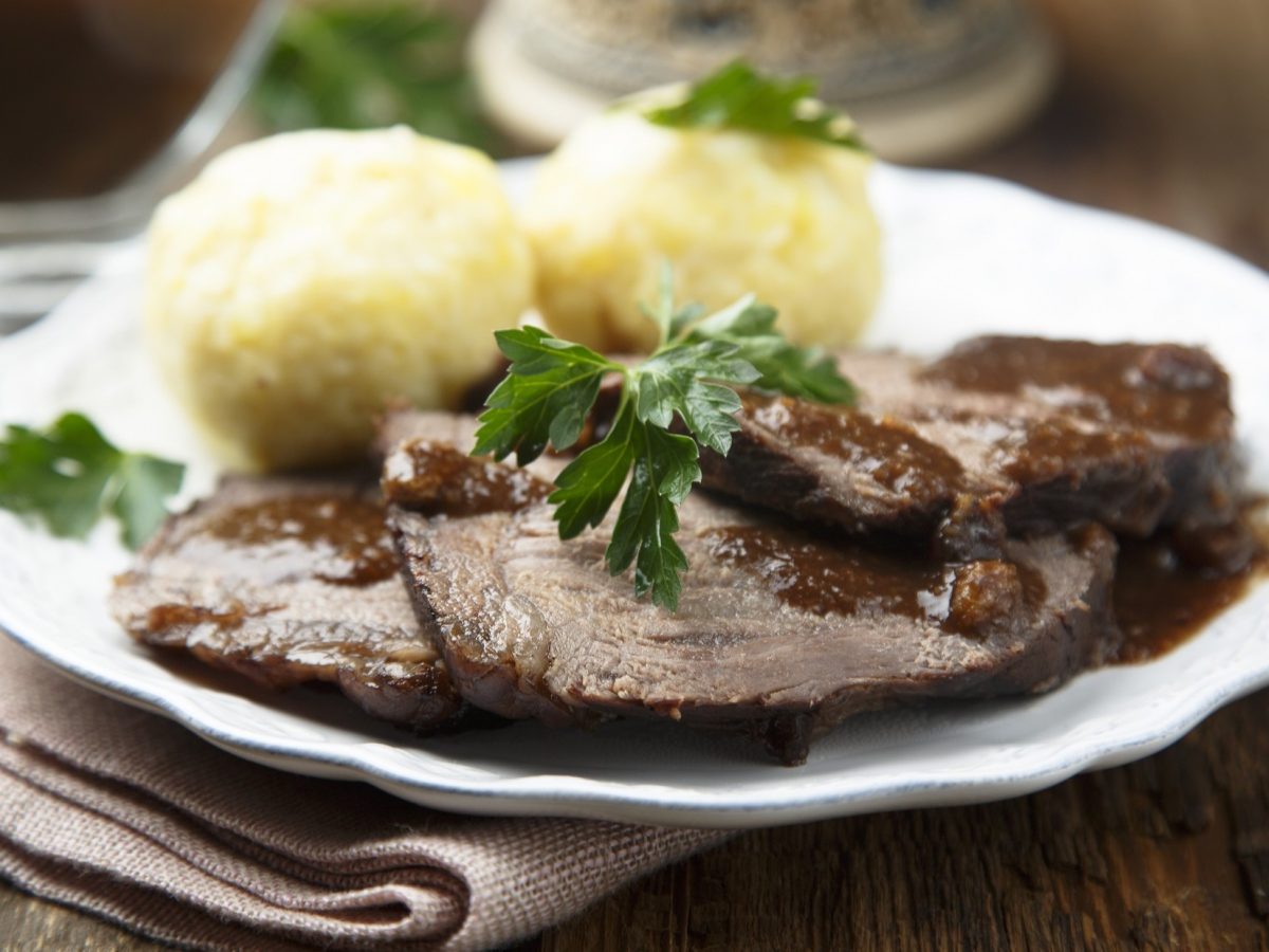 Frontal: Ein Rheinischer Sauerbraten mit zwei Kartoffelklößen auf einem weißen Teller. Das Gericht ist mit frischer Petersilie garniert.