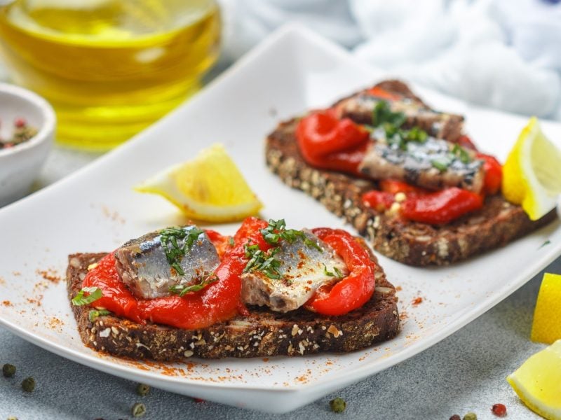 Draufsicht, schräg: Ein weißer, rechteckiger Servierteller mit zwei Sardinen-Crostini mit gerösteter Paprika. Daneben liegen mehrere Zitriónenschnitzer.