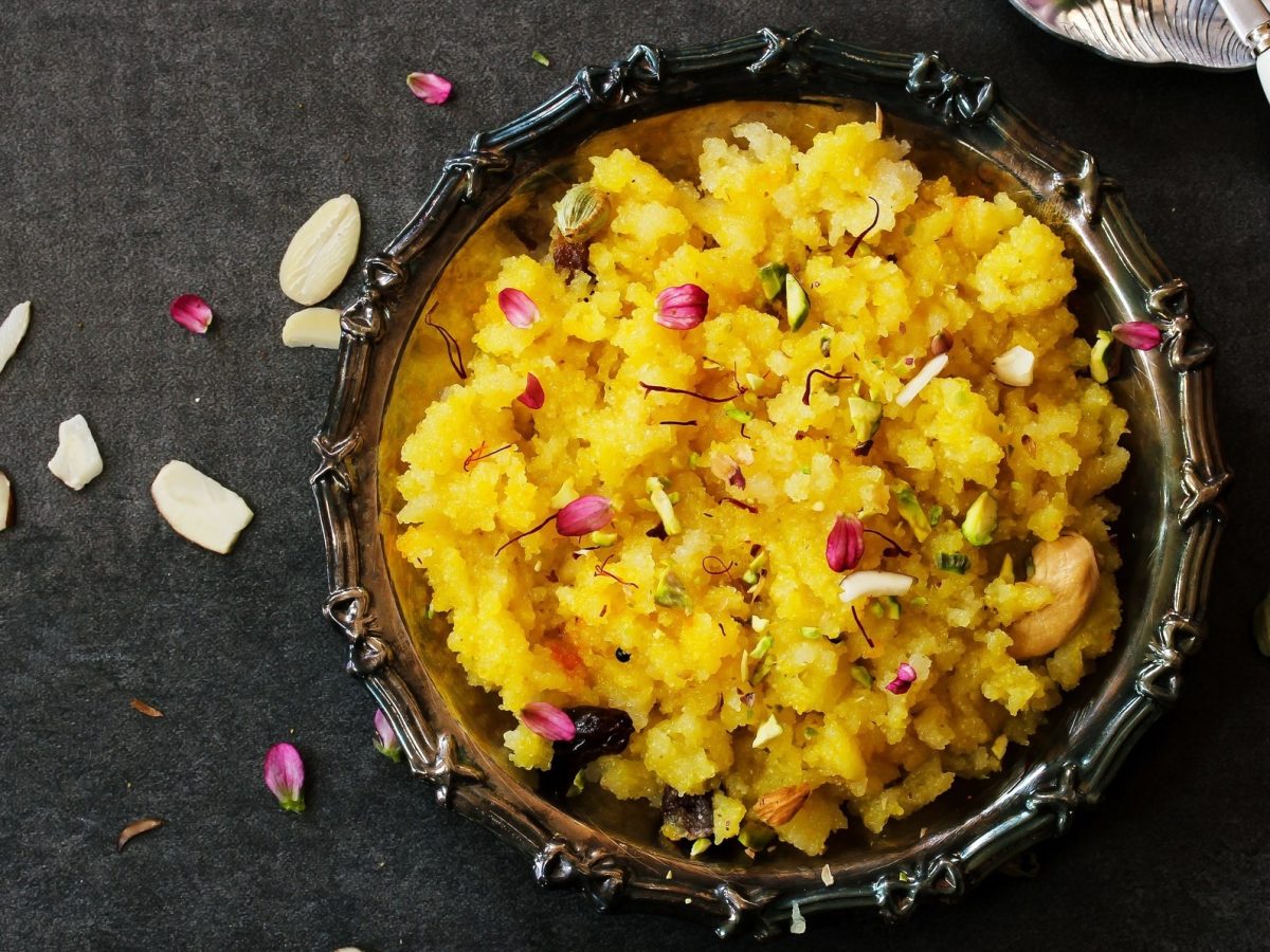 Draufsicht: Eine verzierte Schale aus Metall mit dem indischen Dessert Sooji ka halwa. Die steht auf einem dunklen Untergrund. Drum herum sind Mandeln und Rosenblätter verstreut.