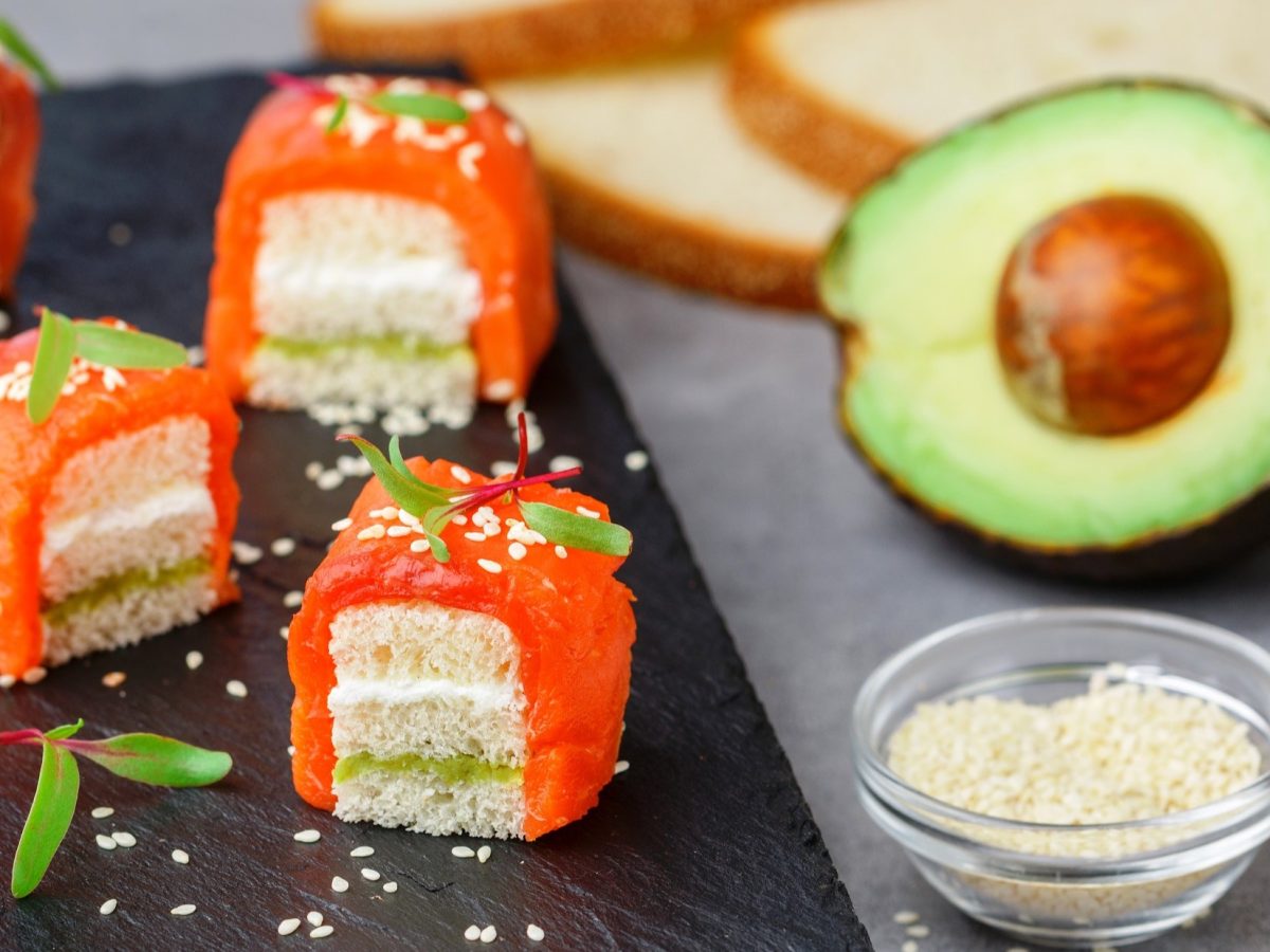 Draufsicht, schräg: Mehrere Sushi-Würfel mit Lachs liegen auf einem dunklen und einen grauen Untergrund. Daneben ist ein Schälchen mit Lachs, eine halbe Avocado und mehrere Scheiben Weißbrot.