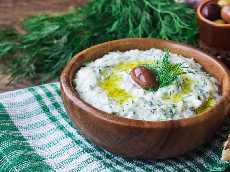 Draufsicht, schräg: Eine Tonschale mit Tsatsiki. Auf dem Dip liegen frischer Dill und ein Olive. Im Hintergrund ist eine weitere Schale mit Oliven und frischer Dill.