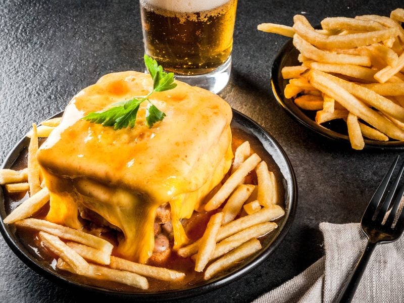 Francesinha auf einem schwarzen teller mit Pommes, daneben eine Schüssel mit Pommes und ein Glas Bier.