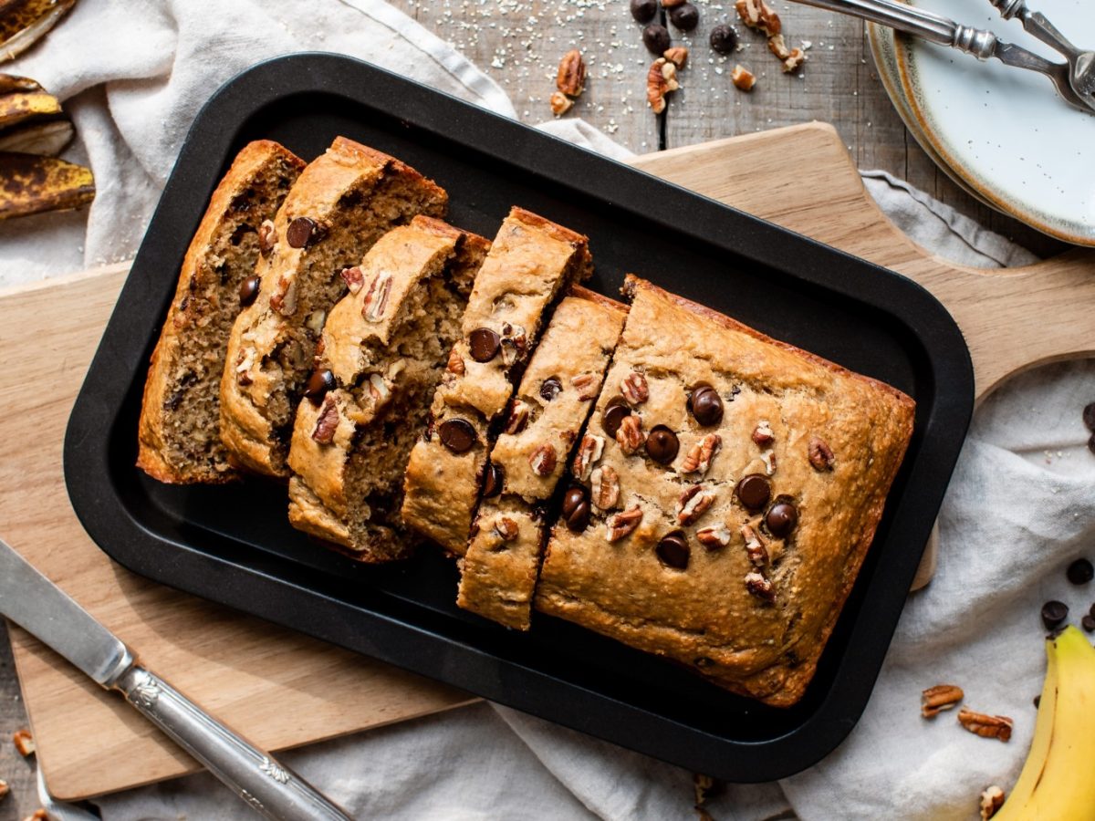 Glutenfreies Bananenbrot auf einem schwarzen Teller auf einem braunen Schneidebrett. Links ein Messer, rechts Bananen und Schokodrops.