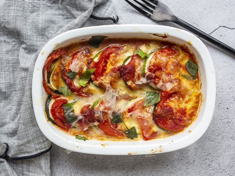 Veganes Tomaten-Kartoffel-Gratin in einer weißen Auflaufform auf grauem Hintergrund. Links ein Geschirrtuch und rechts eine Gabel.