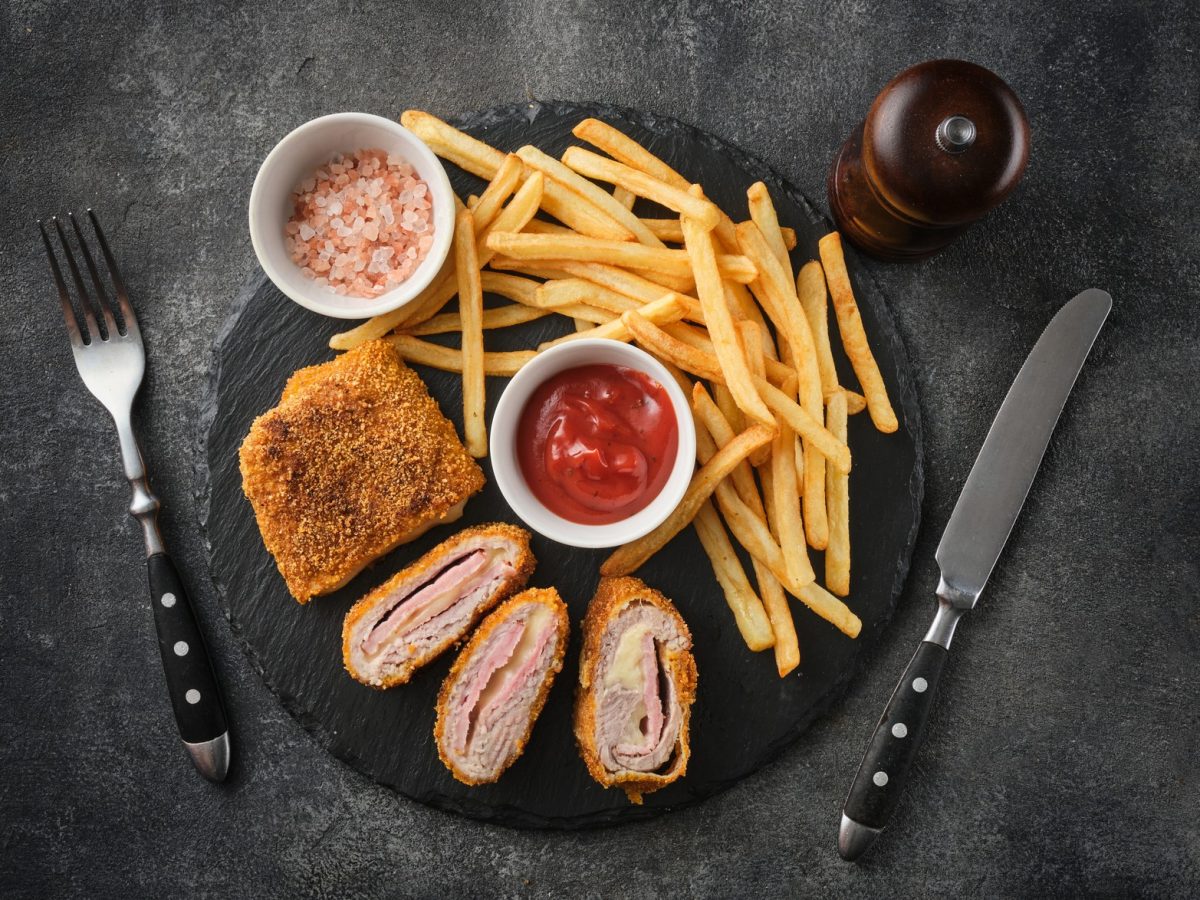 Hähnchen Cordon Bleu, aufgeschnitten, mit sichtbarer Füllung, umgeben von knusprigen Pommes, Ketchup und Gewürzen auf Steinplatte.