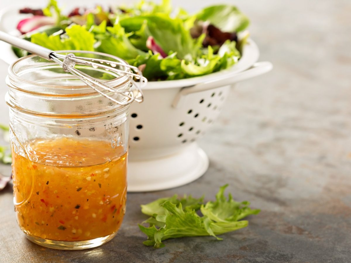 Ein Glas mit dem Italian Dressing auf einem grauen Steintisch. Im Hintergrund eine weiße Salatschüssel mit buntem Salat. Alles von der Seite fotografiert.