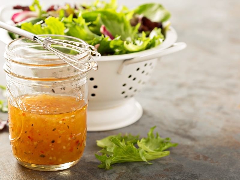 Ein Glas mit dem Italian Dressing auf einem grauen Steintisch. Im Hintergrund eine weiße Salatschüssel mit buntem Salat. Alles von der Seite fotografiert.