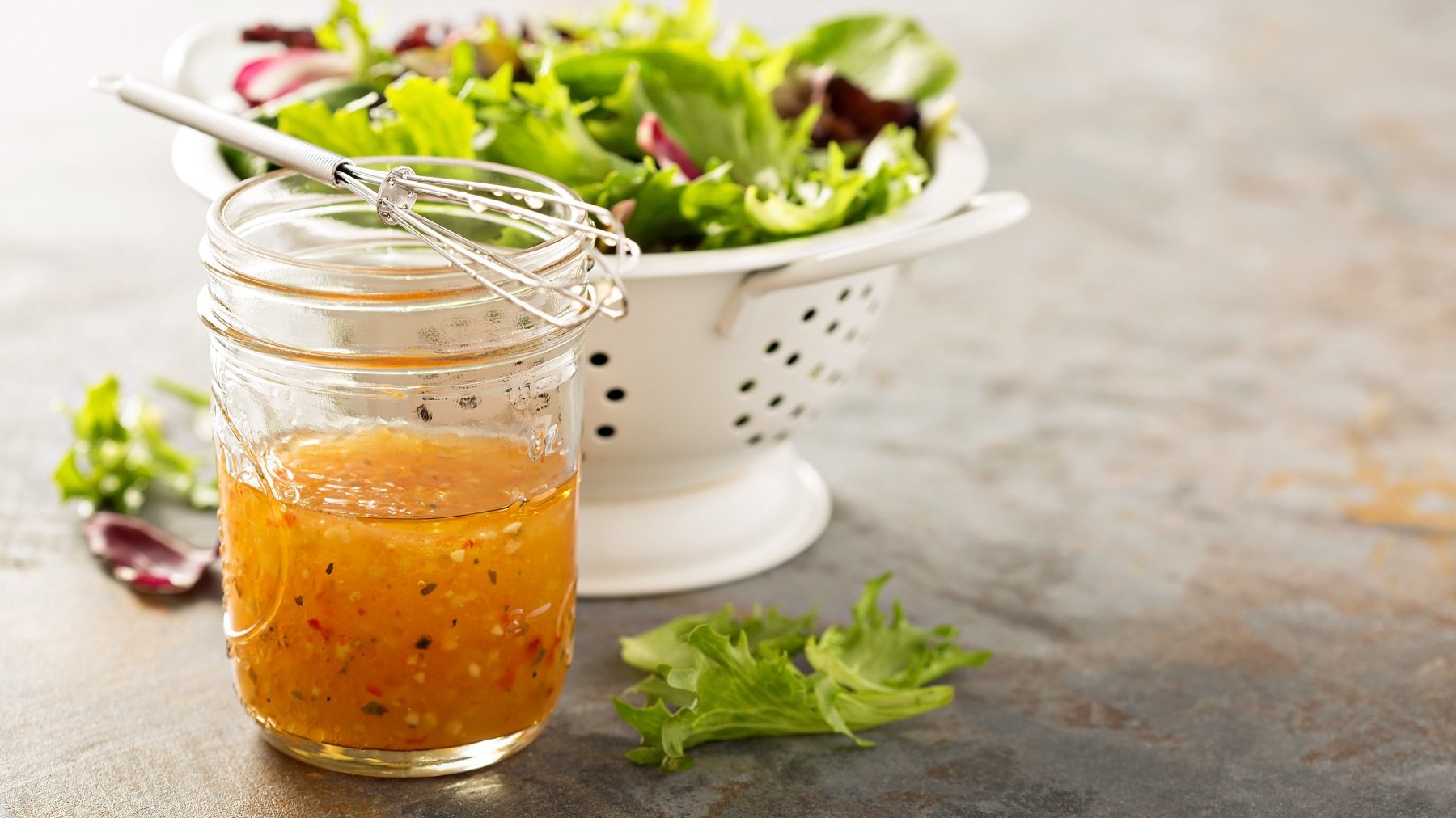 Ein Glas mit dem Italian Dressing auf einem grauen Steintisch. Im Hintergrund eine weiße Salatschüssel mit buntem Salat. Alles von der Seite fotografiert.