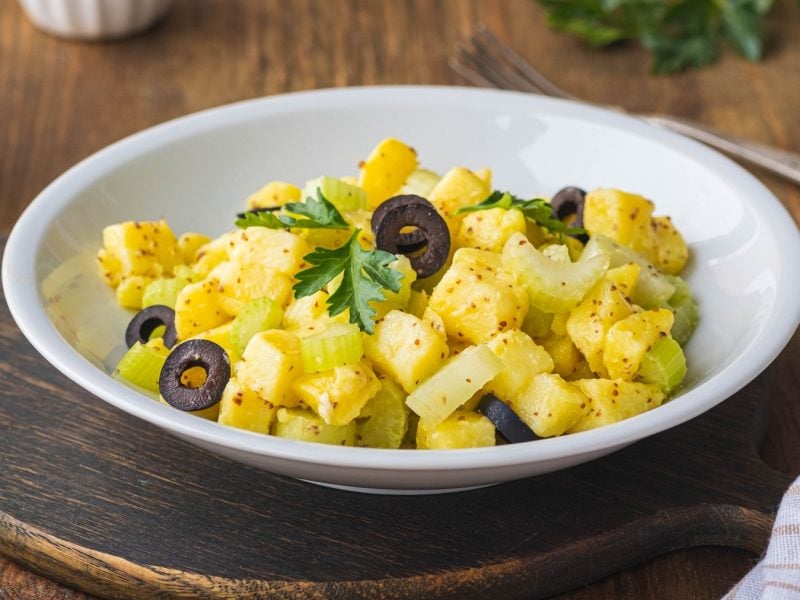 Ein weißer Teller mit dem Kartoffelsalat auf einem rustikalen Holztisch. Im Hintergrund Öl, Petersilie und Holzdeko.