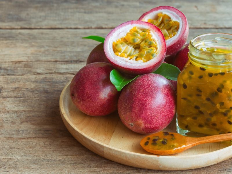 Maracuja-Marmelade in einem Glas auf einem beigen Teller auf braunem Hintergrund. Auf dem Teller sind ebenfalls Maracujas und ein Löffel.