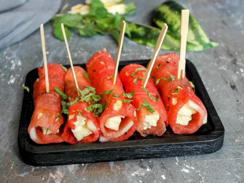 Einige Melonen-Halloumi-Röllchen auf einem kleinen schwarzen Brett und Steintisch, mit Minze bestreut. Im Hintergrund unscharf Wassermelone, Minze und ein graues Tuch zu erkennen.