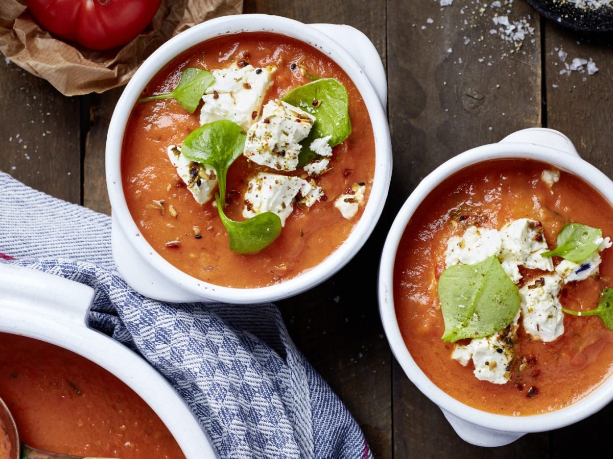 Zwei Schüsseln gefüllt mit Ofentomatensuppe mit mariniertem Deichkäse auf braunem Hintergrund. Links unten ein Topf mit Tomatensuppe und ein blaues Geschirrtuch.