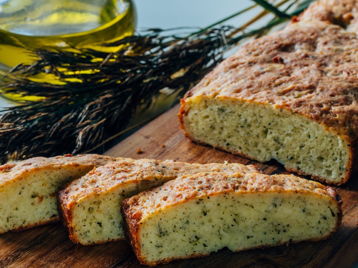 Das Parmesanbrot angeschnitten auf einem rustikalen Holztisch mit einem grünen Strauch und Olivenöl im Hintergrund. Alles nah und von leicht schräg oben fotografiert.