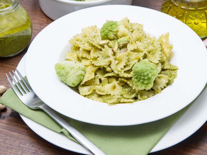 Ein weißer Teller und Unterteller mit einer hellgrünen Serviette und dem Pastagericht. Alles auf einem dunklen Holztisch vom schräg oben fotografiert. Drumherum Zutaten wie das Pesto, Brokkoli, Knoblauch und Olivenöl.