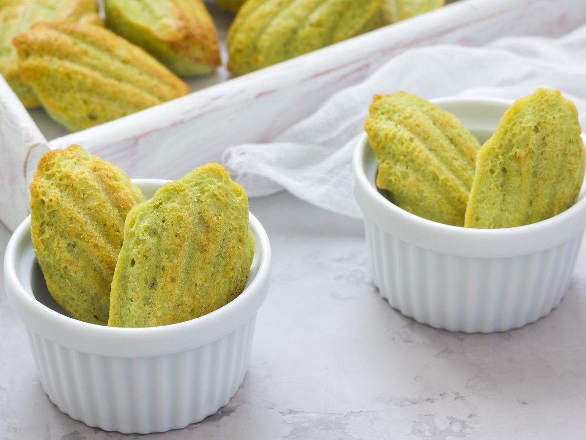 Einige Pistazien-Madeleines in kleinen weißen Schälchen und auf einem großen Brett im Hintergrund. Tisch, Schälchen und Untergrund sind in Grau- und Weißtönen gehalten und von leicht schräg oben fotografiert.