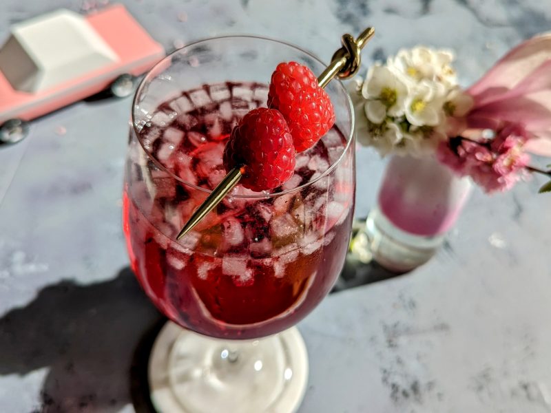 Ein Glas mit dem Cocktail auf grauem Steintisch. Unscharf einige Blüten und ein pinkes Holzauto im Hintergrund zu erkennen.