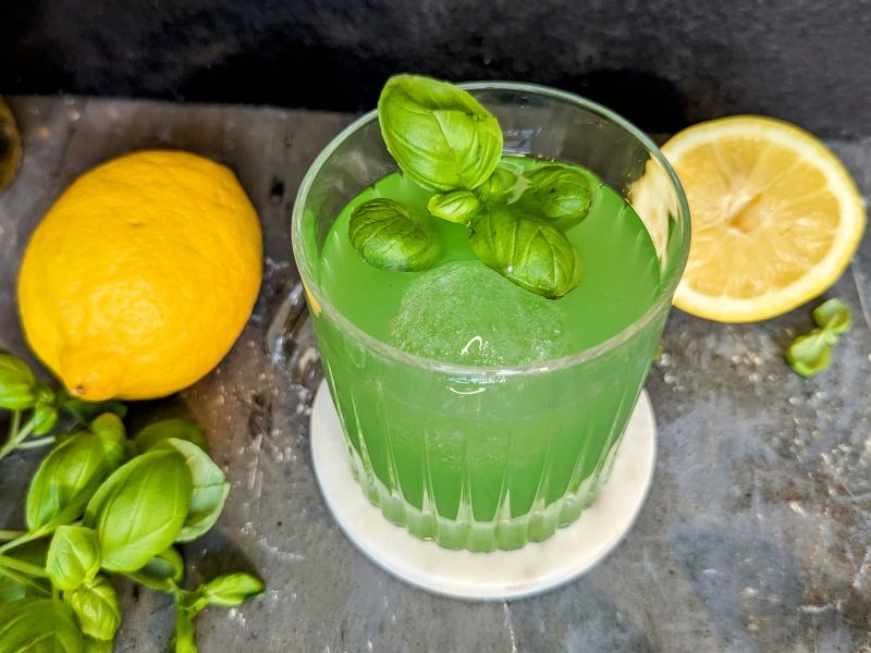 Ein Glas mit dem grünen Cocktail auf einem grauen Steintisch, mit Zitrone, Basilikum und Lillet im Hintergrund.