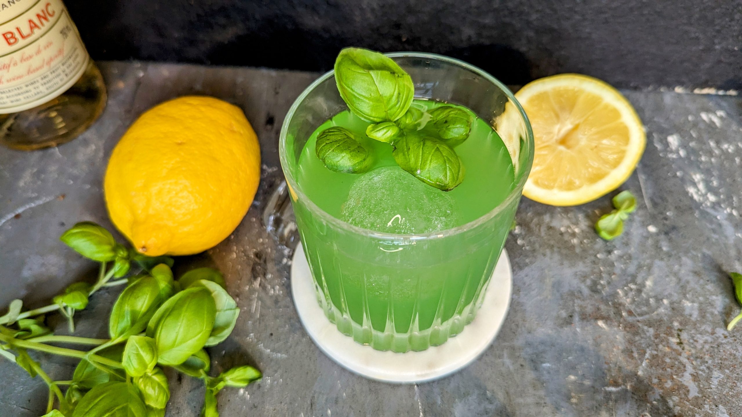 Ein Glas mit dem grünen Cocktail auf einem grauen Steintisch, mit Zitrone, Basilikum und Lillet im Hintergrund.