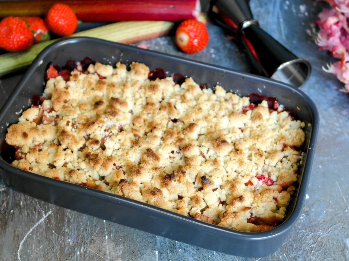 Eine graue Auflaufform mit dem Crumble auf einem grauen Steintisch. Dahinter pinke Blüten und einige Zutaten für das Dessert.