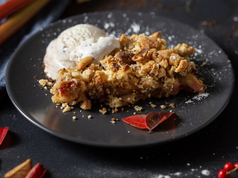 Ein dunkler Teller mit dem Crumble auf einem dunklen Tisch mit roter Deko und Rhabarberstangen.